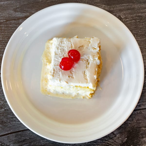 A slice of tres leches cake with whipped cream and two red cherries on a white plate.