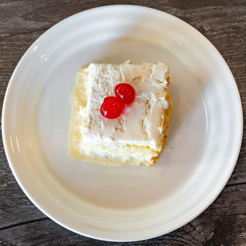 A slice of tres leches cake with whipped cream and two red cherries on a white plate.